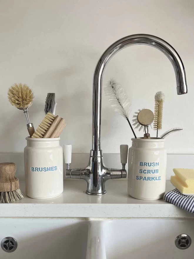   Handmade ‘brushes’ and ‘brush scrub sparkle’ jars are filled with scrubbing brushes, by a sink.