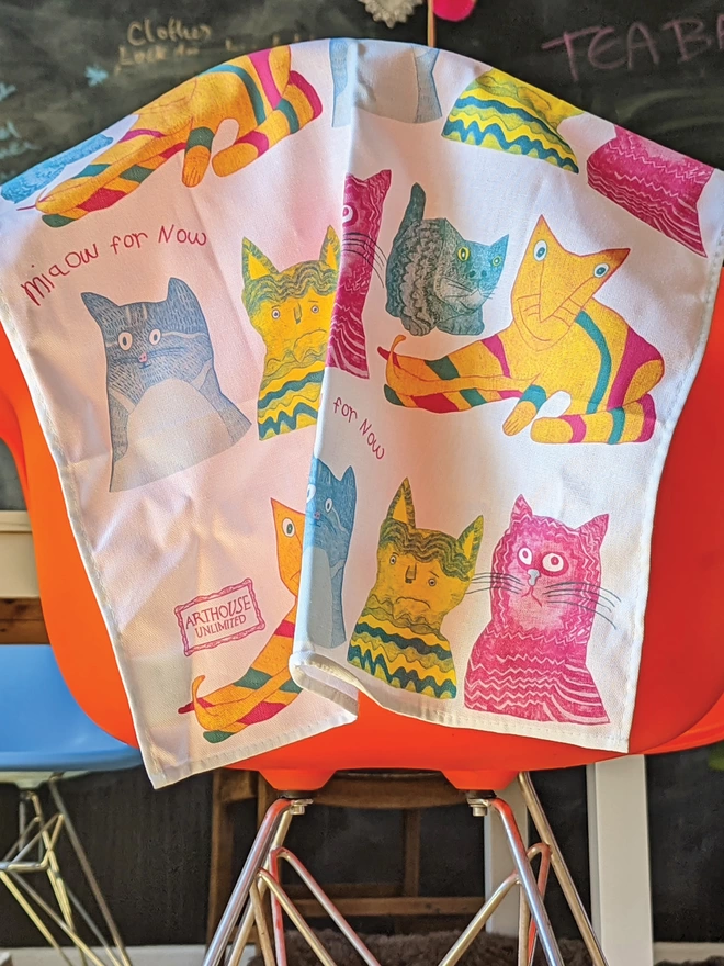 miaow for now 100% organic cotton charity tea towel in blue pink & yellow hanging over an orange chair