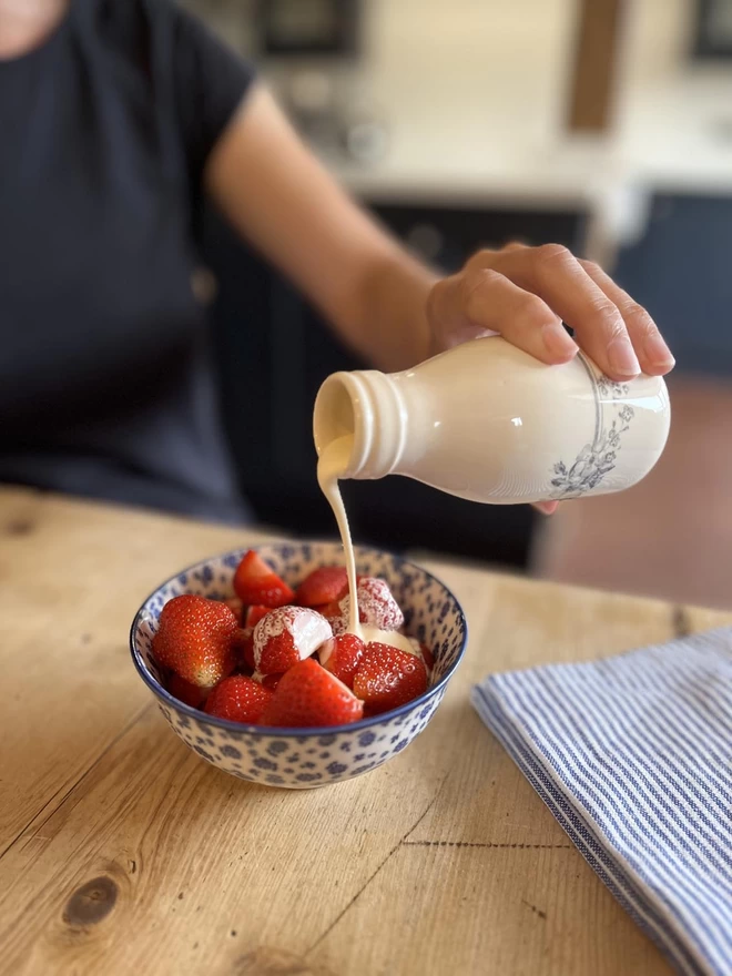  A small handmade ceramic ‘milk’ bottle is pouring cream over a bowl of fresh strawberries.