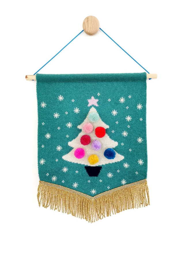 A product image of a jade green knitted wall hanging with a white sparkly christmas tree knitted on the front. The tree is adorned with merry and bright rainbow pom poms and edged with a gold bullion trim. The whole banner is suspended from a wooden wall dot by a bright blue nylon cord.