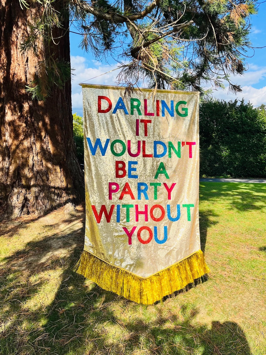 A supersize customisable banner hanging outside. The background is gold sequins and it has a gold tinsel trim along the bottom. The text is mutli-coloured and says 'DARLING IT WOULDN'T BE A PARTY WITHOUT YOU'