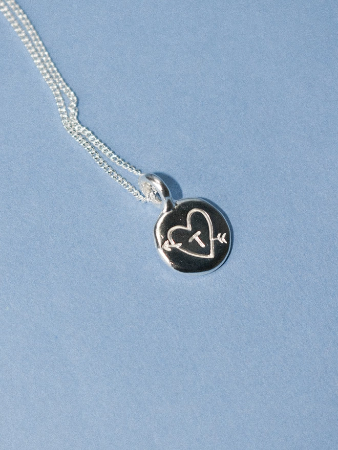 a small silver disk with a hand engraved heart and arrow with a T in the centre. the pendant is attached to a thin silver chain and lays on a pale blue background.