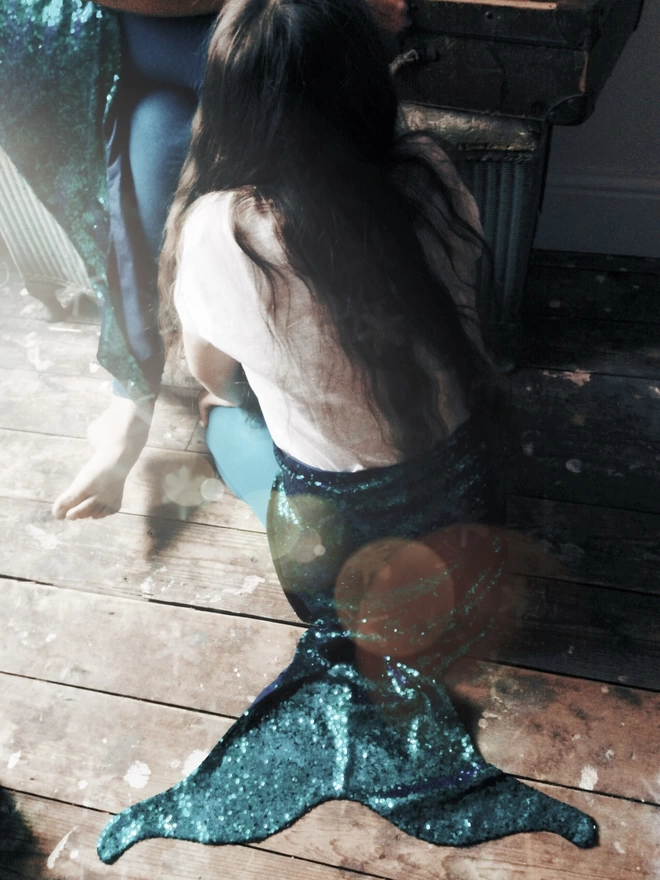The back of a Mermaid with long dark her is kneeling on wooden floorboards.  she is wearing a white t-shirt and a peacock green sequin mermaid tail 