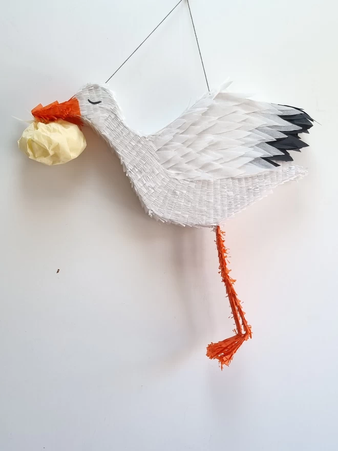 white stalk pinata with feathered wings and an orange beak and legs carrying a small cream bundle in its beak on a white background