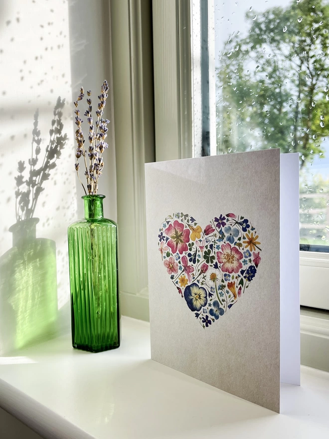 Greetings Card with Digitally Printed Pressed Flower Heart Design Standing on Windowsill - Green Glass Bottle with Dried Lavender - View of Tree and Fence Through Window