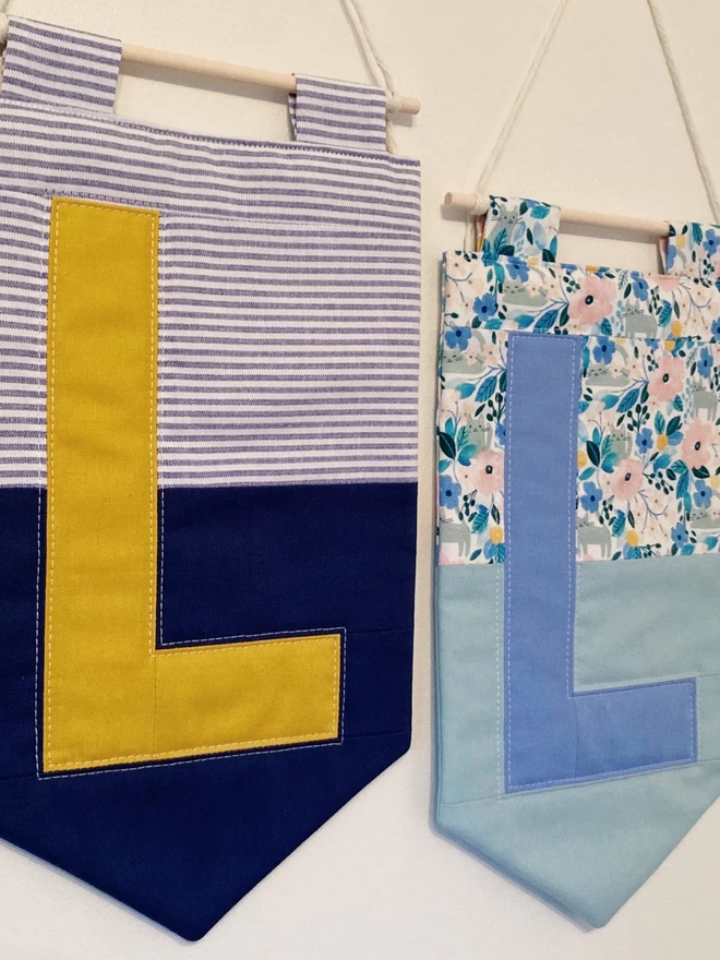 Two Cooper and Fred colour block quilted wall hangings, both with letter 'L', one with stripe and navy background with yellow letter, one with floral and green background with blue letter.