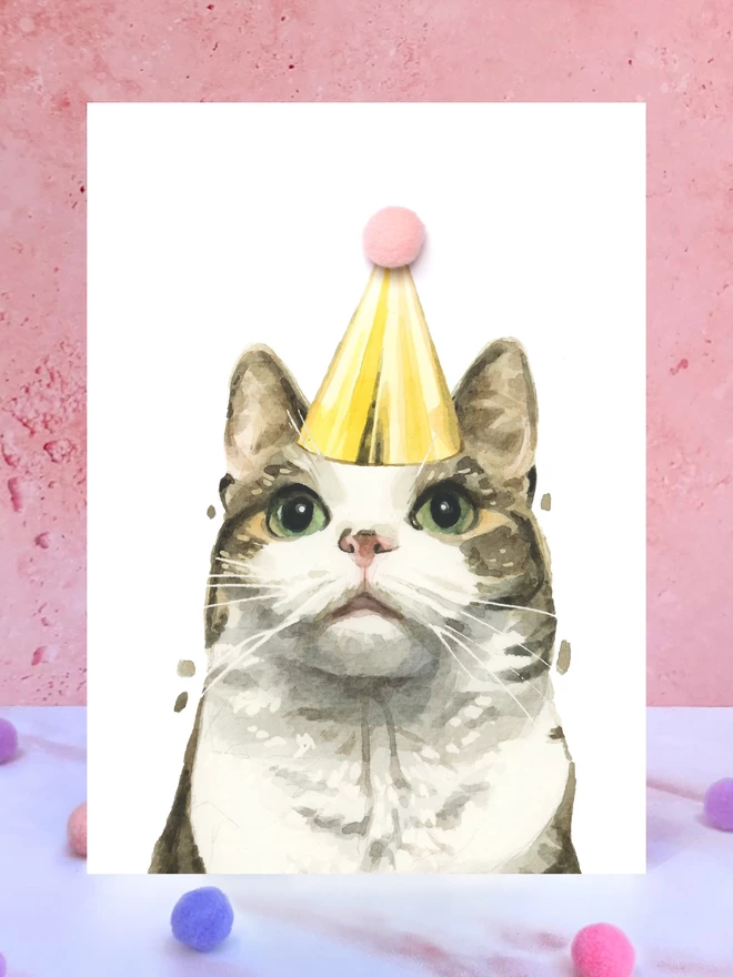 A greeting card featuring a hand painted design of a tabby and white cat, stood upright on a marble surface surrounded by pompoms. 