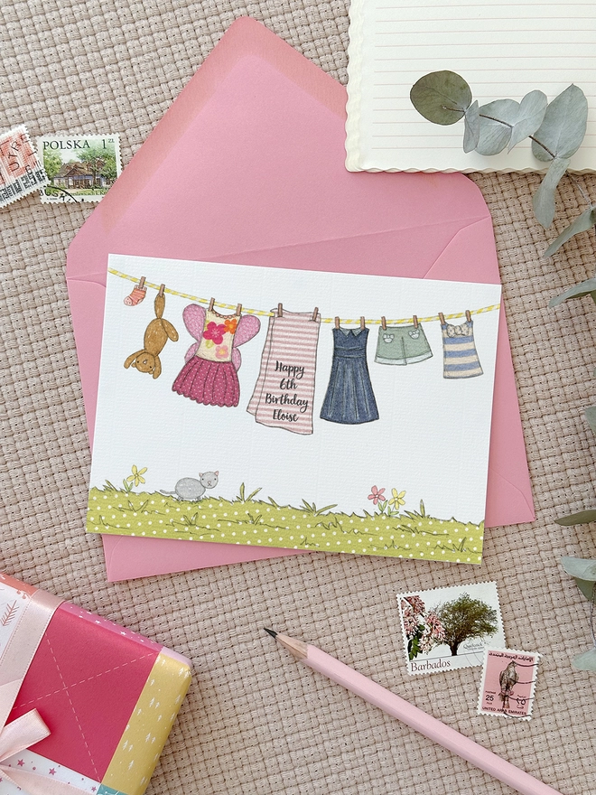 A personalised birthday greetings card with an illustrated washing line of children's clothes lays on a pink envelope beside various stationery items.