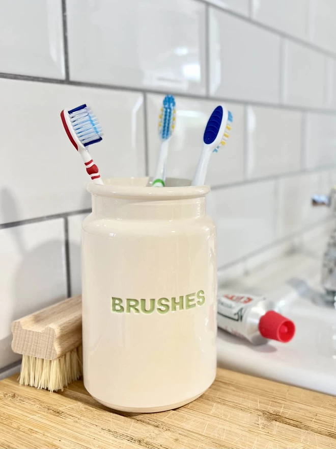 A handmade ‘brushes’ jar (lettering in green) is being used to hold toothbrushes.