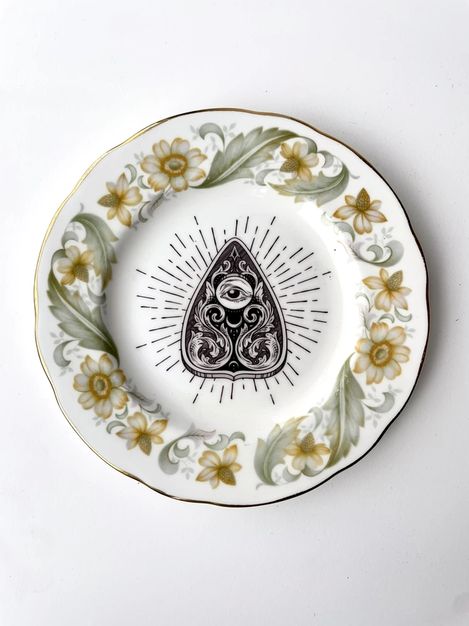 vintage plate with an ornate border, with a printed vintage illustration of a Ouija planchette in the middle 