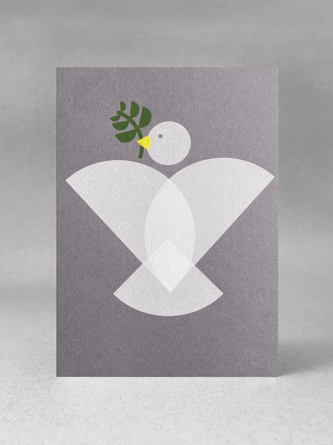 Geometric shapes make up a dove design on this handprinted christmas card, with an olive branch and gold beak. Stood front on in a studio set.