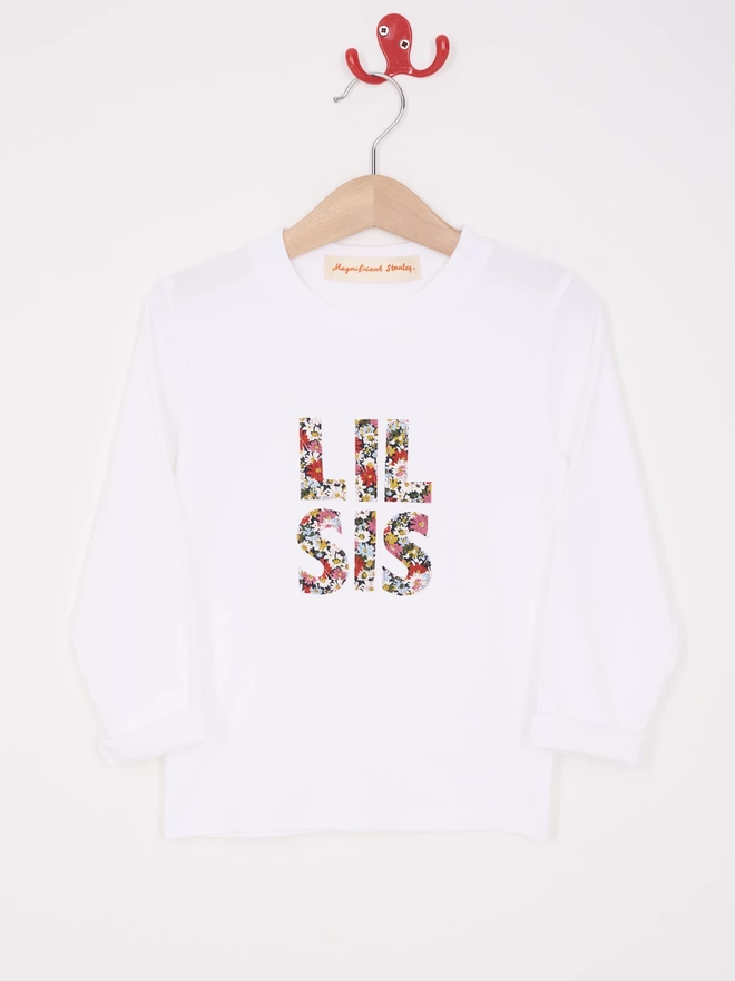 Lil Sis Appliquéd on a white long sleeve cotton white t-shirt in a floral Liberty print, hanging on a hanger