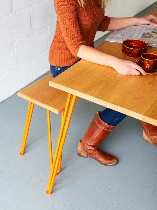 woman sat drinking a coffee at a hairpin leg dining table and bench set with oak tops and orange legs