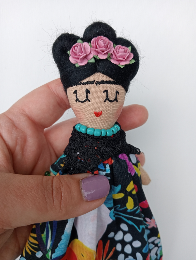 Frida Kahlo mini decorative doll held in a hand against a white backdrop Frida wears a floral dress and paper flower crown