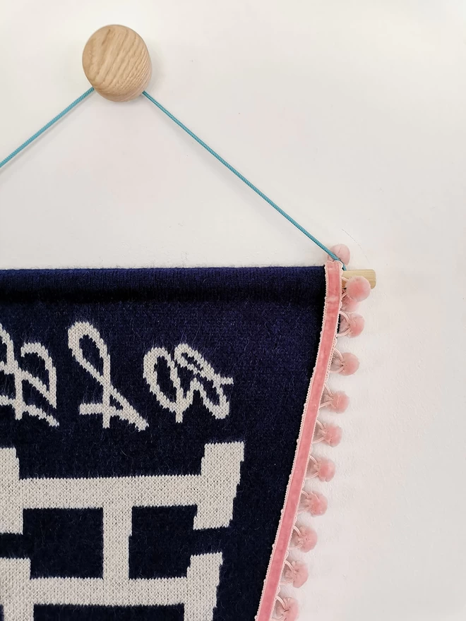 A close up product image of the front of a navy knitted pennant flag wall hanging. The start of the word ‘Bah Humbug’ can be knitted in white lambswool with a hint of gold sparkle in the text.You can see the letters ‘BAH H’ here in detail alongside the blush pink pom pom trim, wooden dowelling and bright teal pom pom cord.