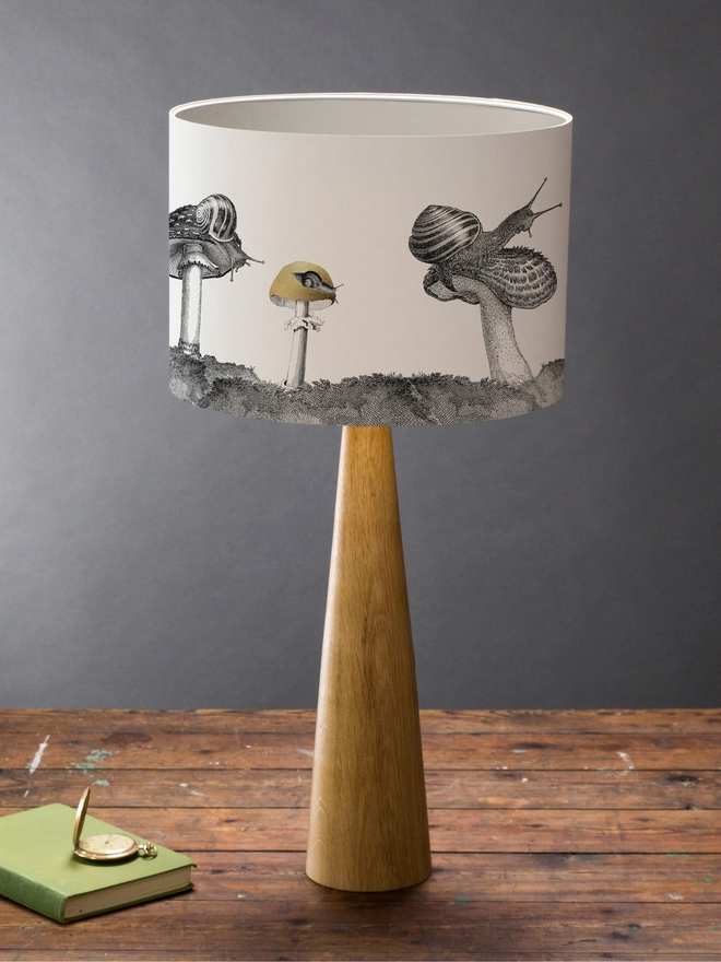 Drum Lampshade featuring snails sitting on mushrooms and toadstools on a wooden base on a shelf with books and ornaments