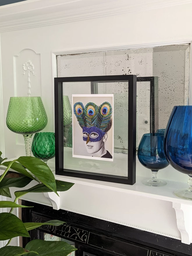 Print of woman wearing embroidered peacock mask framed on shelf