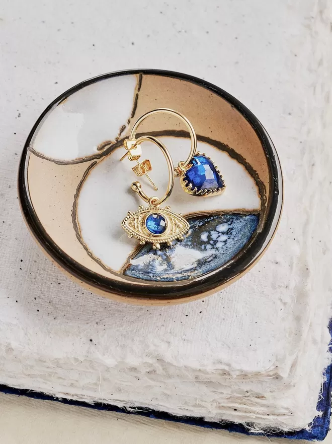 Mismatching gold hoop earrings with blue evil eye charm and blue faceted heart charm on gold background 