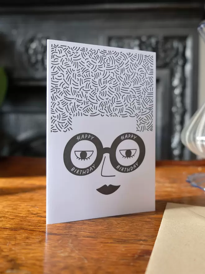 White card with a ladies face wearing glasses saying ' Happy Birthday' and big square hair. 