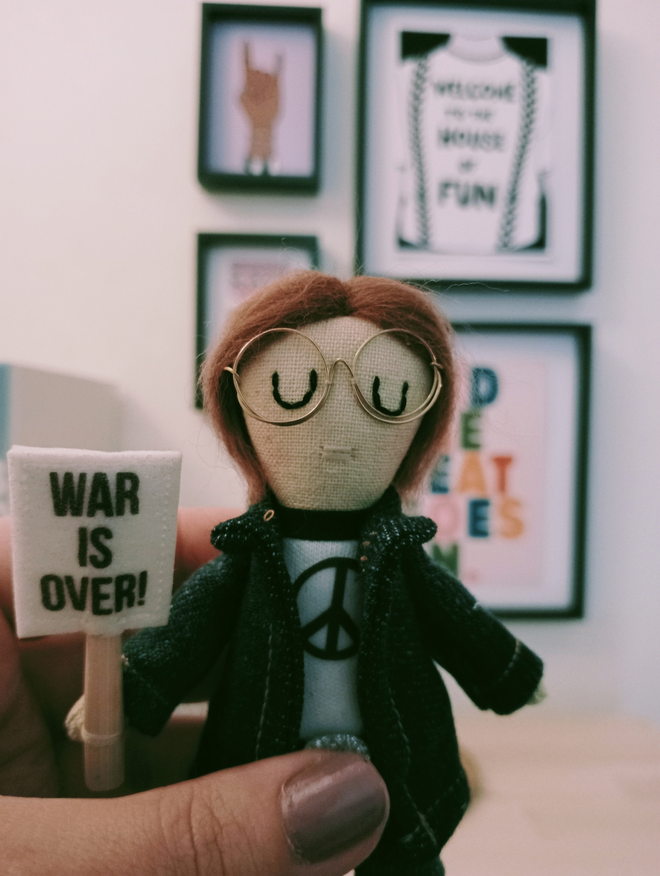 Decorative mini icon doll of John Lennon held in a left hand in front of a small gallery wall of music themed prints John is wearing a denim over shirt and white peace sign emblazoned t shirt in his right hand is a placard bearing the words war is over 