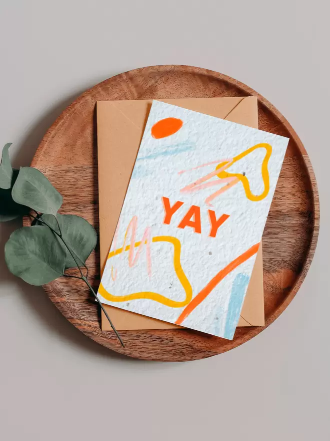 Plantable Card with ‘YAY’ in the centre of the card with an abstract pattern around it on a wooden tray next to a Eucalyptus branch