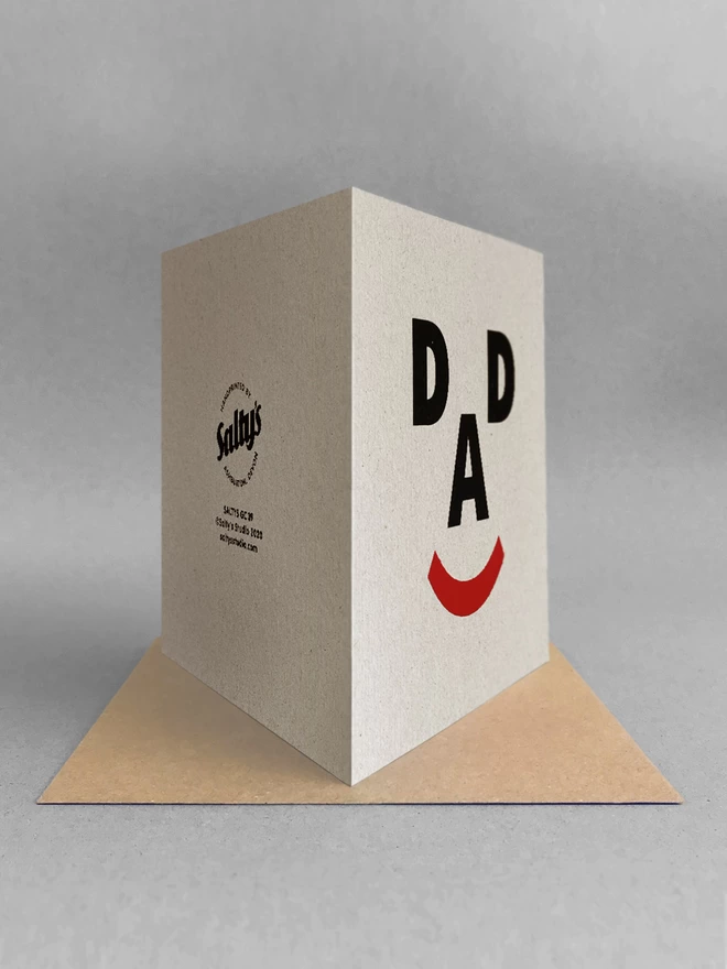 Rear view of a greetings card, the letters D, A, D and a closed bracket symbol make up a happy face, screenprinted on recycled grey card. Stood on a kraft brown envelope, in a light grey studio set
