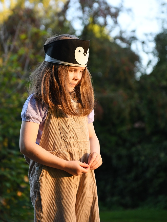 A young girl wearing beige dungarees and a handmade penguin fabric crown stands outside at sunset.