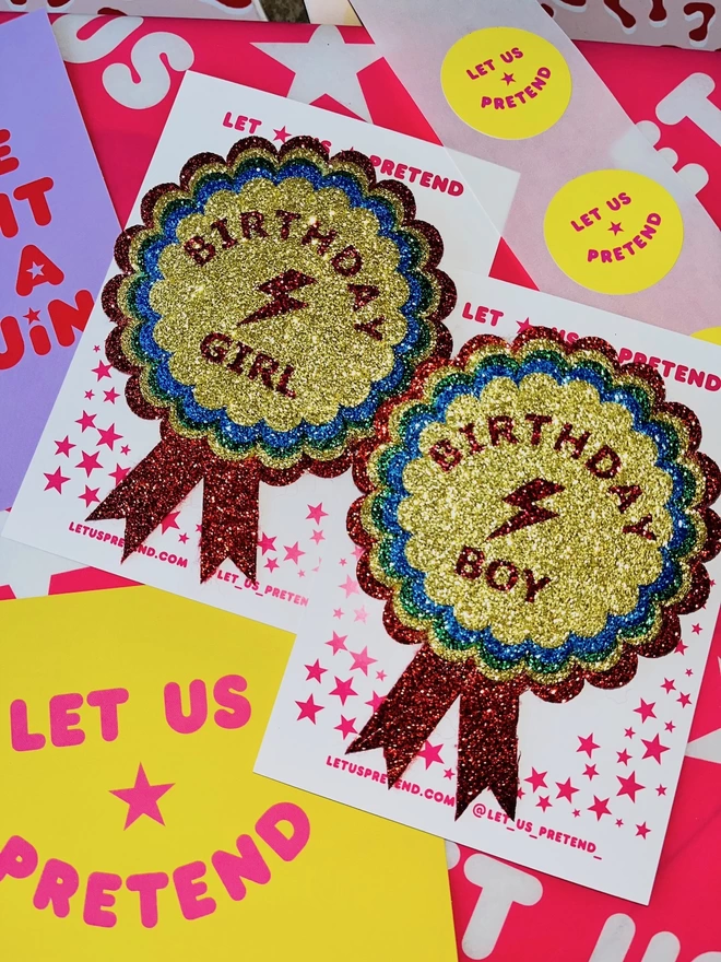 A close up of the 'BIRTHDAY GIRL' and 'BIRTHDAY BOY' badges. In the centre of each badge is a red lightning bolt between the text. The text is on top of layers of scalloped circles in the colours gold, blue, green, and red.