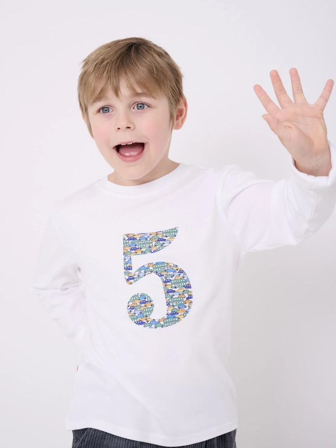 A 5 year old boy wearing a white cotton long sleeve t-shirt appliquéd with the number 5  in Liberty print featuring vintage cars and busses