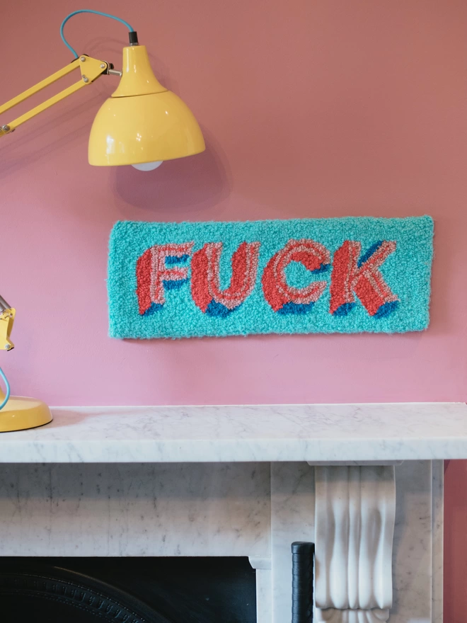'FUCK' Handmade Tufted Rug/Wall Hanging seen in blue hanging on a pink wall with a yellow light.