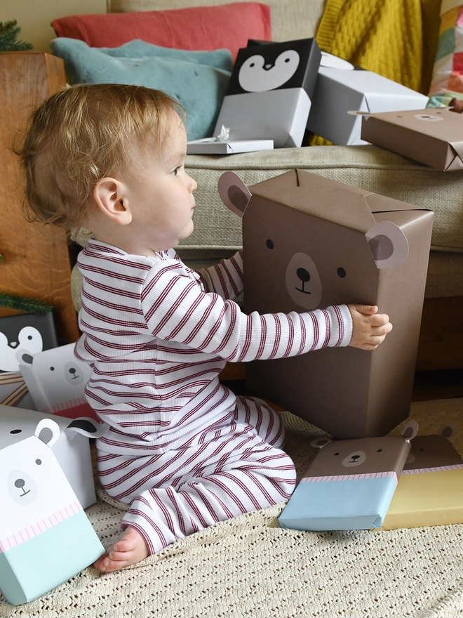 A baby wearing red and white striped pyjamas holds a gift wrapped as brown bear and is surrounded by other gifts wrapped in animal wrapping paper.