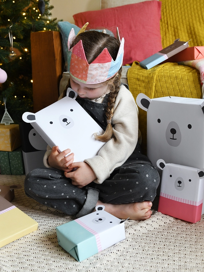 A young girl wearing grey dungarees and a patchwork crown holds a gift wrapped as polar bear and is surrounded by other gifts wrapped in animal wrapping paper.
