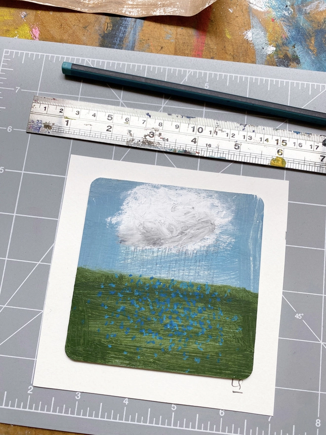 painting of blue sky, a cloud and a field of blue forget-me-nots on cutting board with steel ruler 