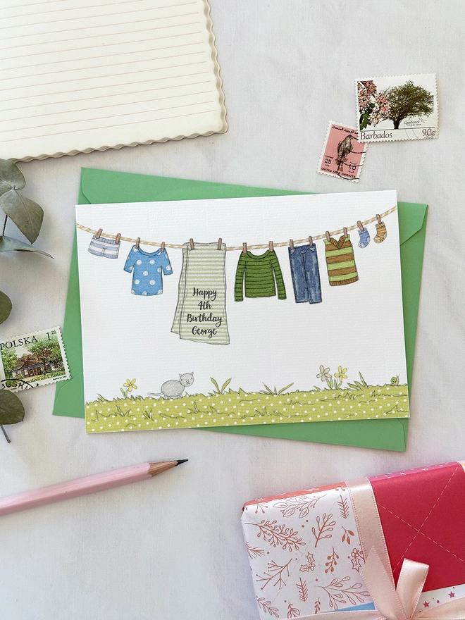 A personalised birthday greetings card with an illustrated washing line of children's clothes lays on a green envelope on a white desk.