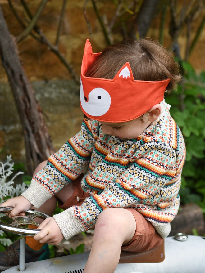 A young child wearing a knitted jumper and a fox dress up fabric crown sits on a ride on vintage car in the garden.