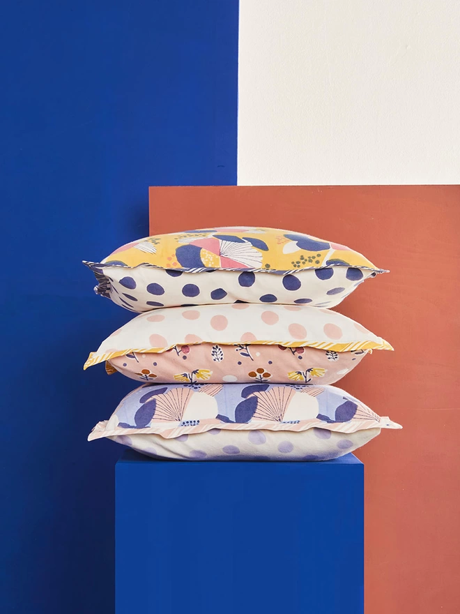 Pile of three block printed cushions, one yellow and navy, one pink and yellow in the middle and the bottom one in shades of blue placed ion a bright blue plinth in front of a blue background
