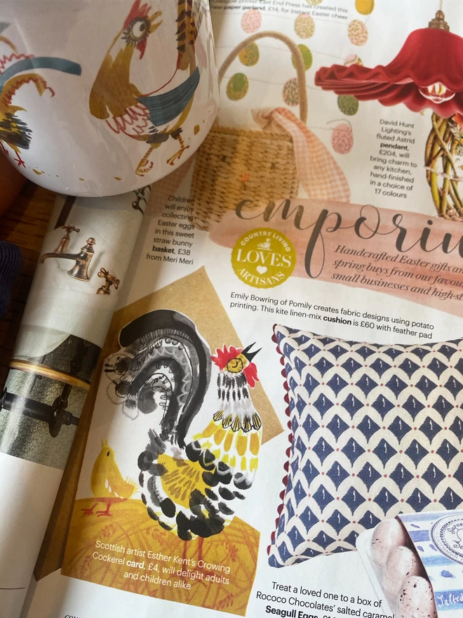 Shaped Crowing Cockerel greetings card illustrated by Esther Kent in in yellow and black, is shown in Country Living Magazine spread. Cockerels Enamel Mug is partly visible.