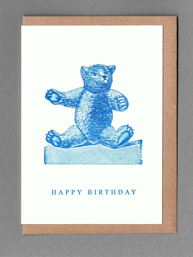 White card with blue illustration of a teddy bear and text reading 'Happy Birthday' with a brown envelope behind
