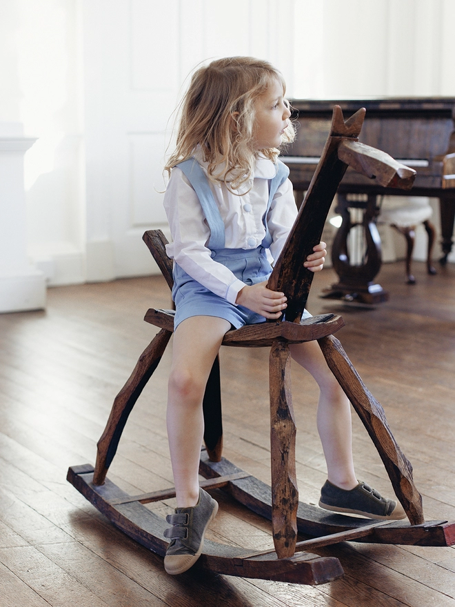 A little boy sits on a rocking horse wearing a white peter pan  collared shirt and blue shorts with braces