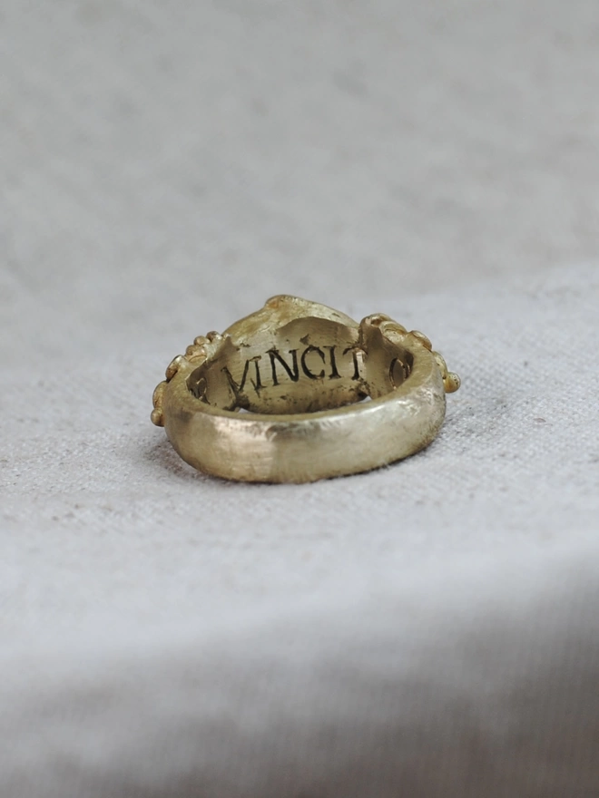 The image of a gold toned brass ring having been turned so the top of the ring is facing away from the camera and the top middle of the inside of the band is showing, revealing the engraved Latin word vincit.