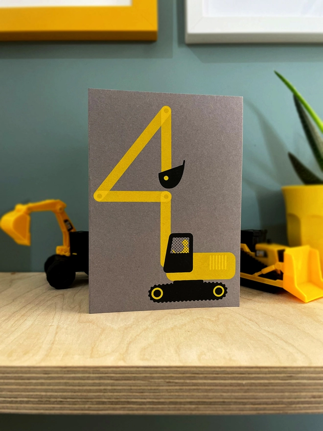 A yellow and black digger, with its arm bent into a number 4 shape - screenprinted onto a grey card. Its is stood on a plywood shelf with a digger toy in the background. some picture frames around the edges and a plant half in shot.