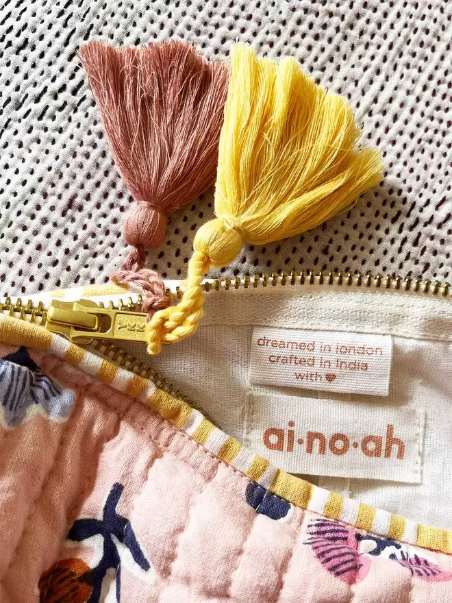 Close up of ainoah label, gold zip and two tassels, one yellow and one soft pink 