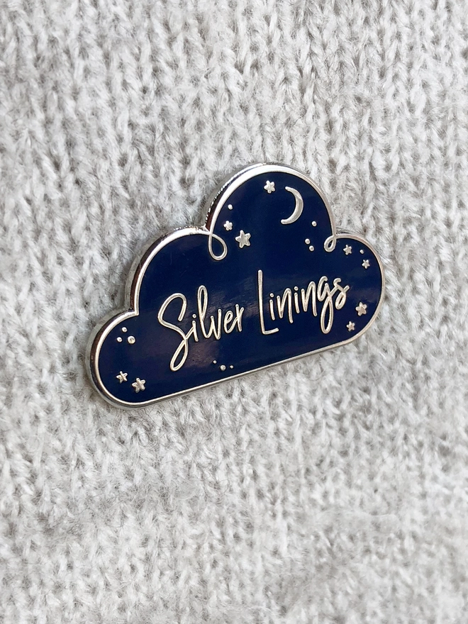 A navy blue and silver enamel pin badge in the shape of a cloud with a starry design and the words "Silver Linings" is pinned to a grey jumper.