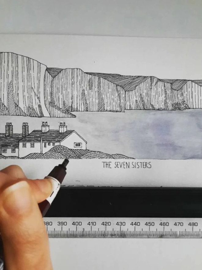Artist Katherine Jones using a pen to hand-draw Seven Sisters cliffs and cottage