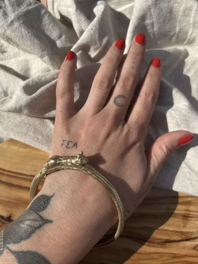 The image of a tattooed hand laying on a piece of raw linen and some rustic wood. The hand has red nail polish and is wearing a gold toned brass bangle with a hand carved tiger on it 