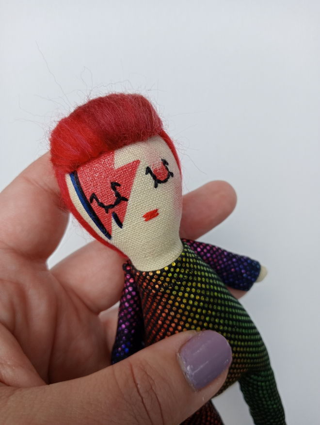 Close up detail of David Bowie mini decorative doll held in a left hand for scale David is wearing a rainbow hued metallic jumpsuit 