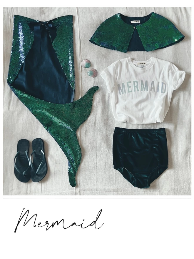 a square flat-lay of a mermaid costume. A long peacock green sequin mermaid tail is positioned on the left the tail flicked up to reveal flip flops. On the top right is a peacock green sequin cape, middle right a white t-shirt with a mermaid slogan in an iridescent pearl print, bottom left - dark green velvet shorts.