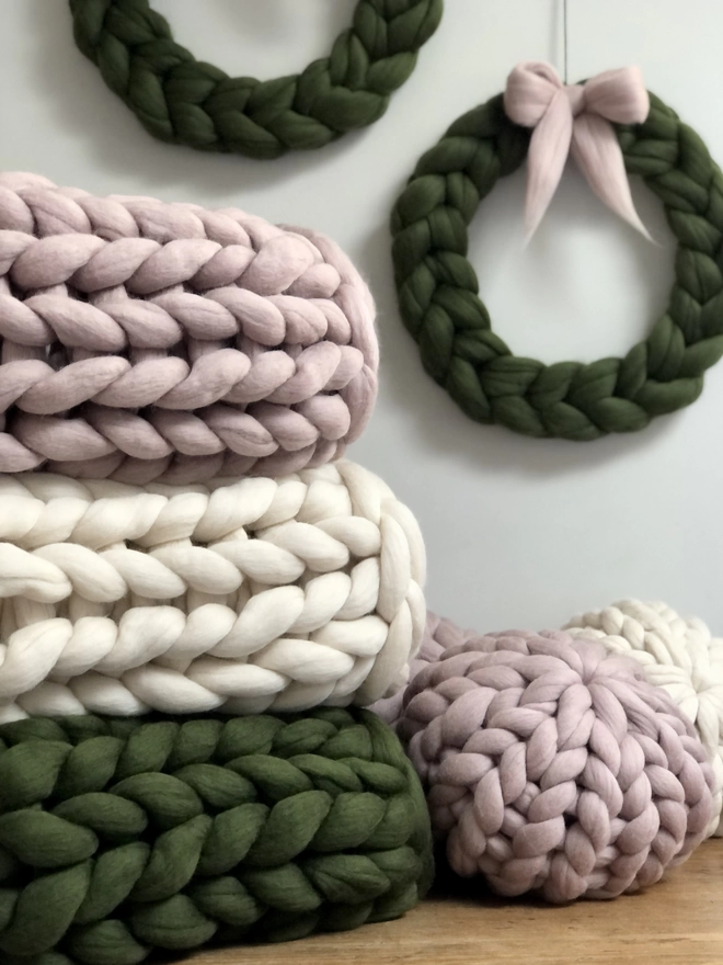 A stack of giant knitted merino blankets in mink pink, oyster and willow green sits on top of an oak blanket box, with two round merino cushions next to it, and two woolly wreaths in willow green are hung on the wall behind