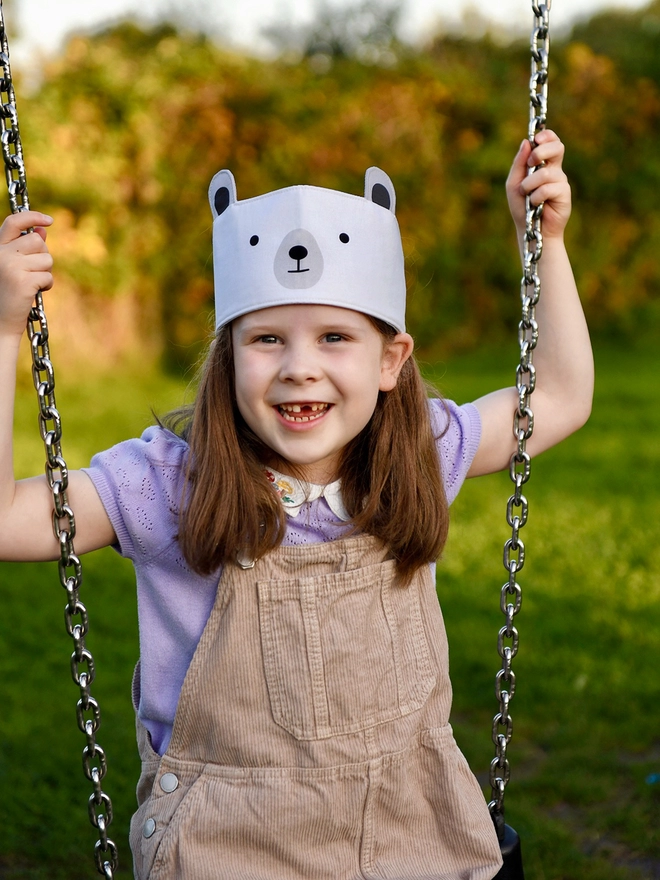 A young girl wearing a dress-up polar bear fabric crown and beige dungarees is sat on a park swing.