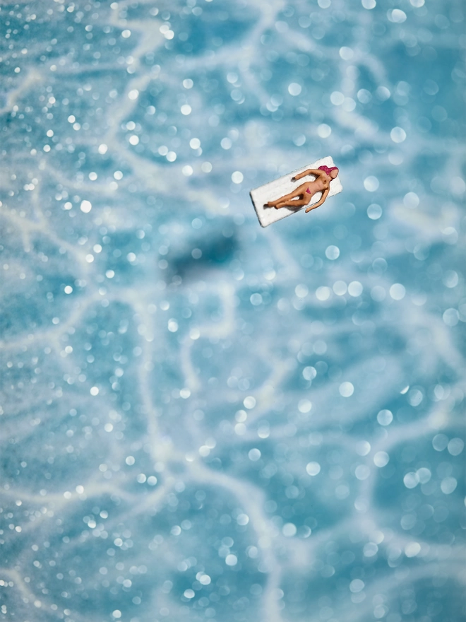 Miniature scene in an artbox showing a tiny naked sunbathing figurine on a lilo, floating on the glass on top of an artbox which looks like a sparkling turquoise swimming pool